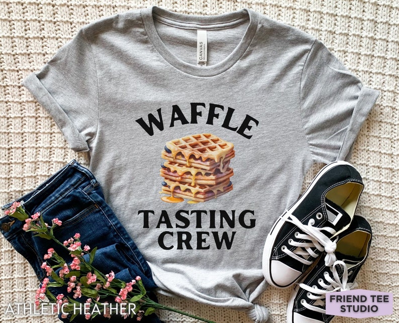 May include: A heather gray t-shirt with the words "WAFFLE TASTING CREW" above a stack of waffles with syrup. The shirt is paired with blue jeans, black sneakers, and a floral arrangement.