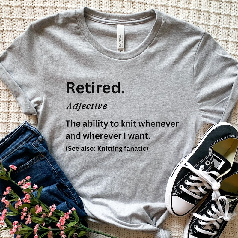 Puede incluir: Camiseta gris jaspeado con el texto "Retired. Adjective. The ability to knit whenever and wherever I want. (See also: Knitting fanatic)". La camiseta se muestra con vaqueros azules y zapatillas negras.