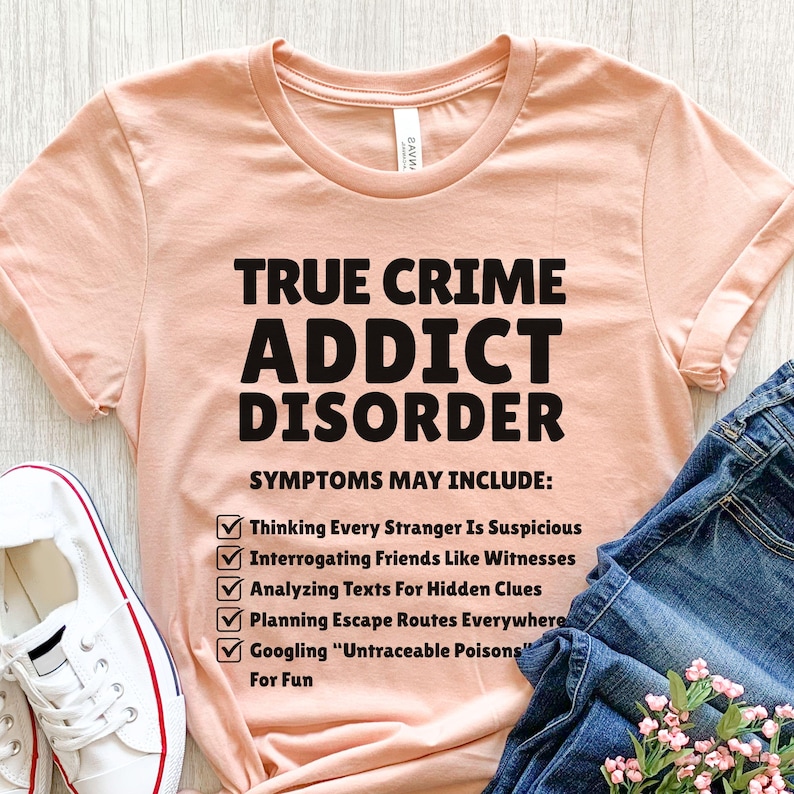 May include: A peach-coloured t-shirt with the text "TRUE CRIME ADDICT DISORDER" in bold black letters. Below, a list of symptoms includes "Thinking Every Stranger Is Suspicious" and "Googling 'Untraceable Poisons' For Fun."