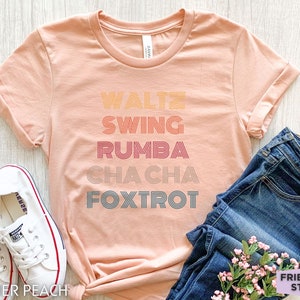 May include: Heather peach-colored t-shirt with the words "Waltz, Swing, Rumba, Cha Cha, Foxtrot" in a retro font. The shirt is paired with blue jeans and white sneakers. The text "Friend Tee Studio" is visible.