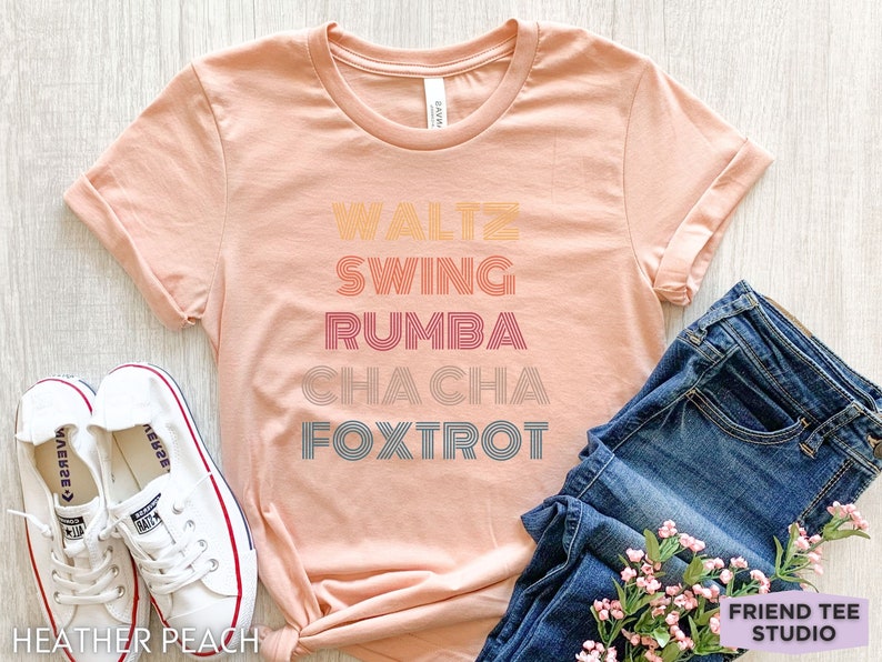 May include: Heather peach-colored t-shirt with the words "Waltz, Swing, Rumba, Cha Cha, Foxtrot" in a retro font. The shirt is paired with blue jeans and white sneakers. The text "Friend Tee Studio" is visible.