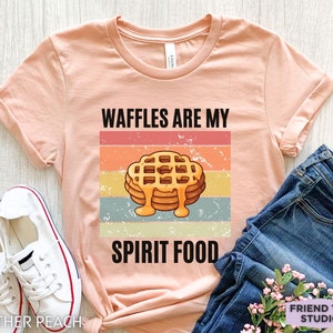 Pu&ograve; includere: T-shirt color pesca con maniche arrotolate, con una grafica di una pila di waffle con sciroppo e la scritta "WAFFLES ARE MY SPIRIT FOOD". Il design &egrave; su uno sfondo a strisce retr&ograve;.