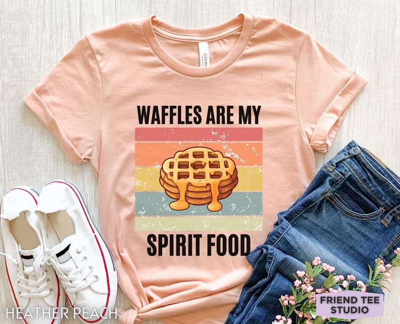 Pu&ograve; includere: T-shirt color pesca con maniche arrotolate, con una grafica di una pila di waffle con sciroppo e la scritta "WAFFLES ARE MY SPIRIT FOOD". Il design &egrave; su uno sfondo a strisce retr&ograve;.