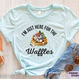 Op de afbeelding: Een lichtblauw T-shirt met de tekst "I'm just here for the waffles" erop gedrukt. De tekst is zwart en er is een afbeelding van een wafel met slagroom, fruit en chocoladesiroop.