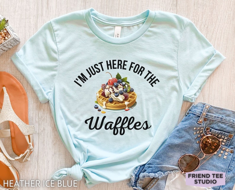 Op de afbeelding: Een lichtblauw T-shirt met de tekst "I'm just here for the waffles" erop gedrukt. De tekst is zwart en er is een afbeelding van een wafel met slagroom, fruit en chocoladesiroop.