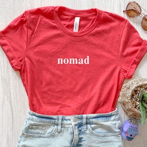 May include: A red t-shirt with the word "nomad" printed in white. The shirt is folded, showing the rolled-up sleeves. The shirt is paired with denim shorts. Sunglasses and a decorative basket are in the background.