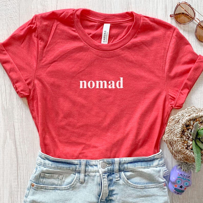 May include: A red t-shirt with the word "nomad" printed in white. The shirt is folded, showing the rolled-up sleeves. The shirt is paired with denim shorts. Sunglasses and a decorative basket are in the background.
