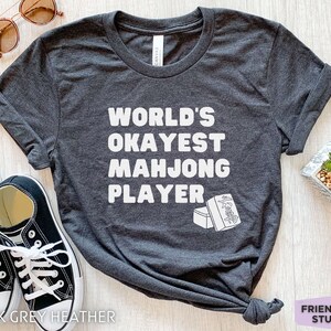 May include: Dark grey heather t-shirt with white text that reads "World's Okayest Mahjong Player" and a graphic of two mahjong tiles.