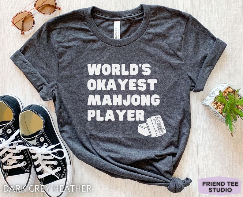 May include: Dark grey heather t-shirt with white text that reads "World's Okayest Mahjong Player" and a graphic of two mahjong tiles.