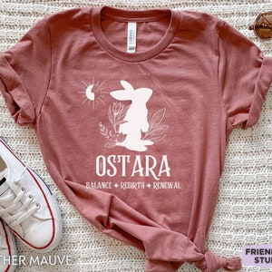 Puede incluir: Una camiseta de manga corta en color malva jaspeado con un gr&aacute;fico blanco de un conejo, flores y un s&iacute;mbolo de sol y luna. El texto dice OSTARA, seguido de las palabras BALANCE, REBIRTH y RENEWAL separadas por peque&ntilde;as estrellas de cuatro puntas.