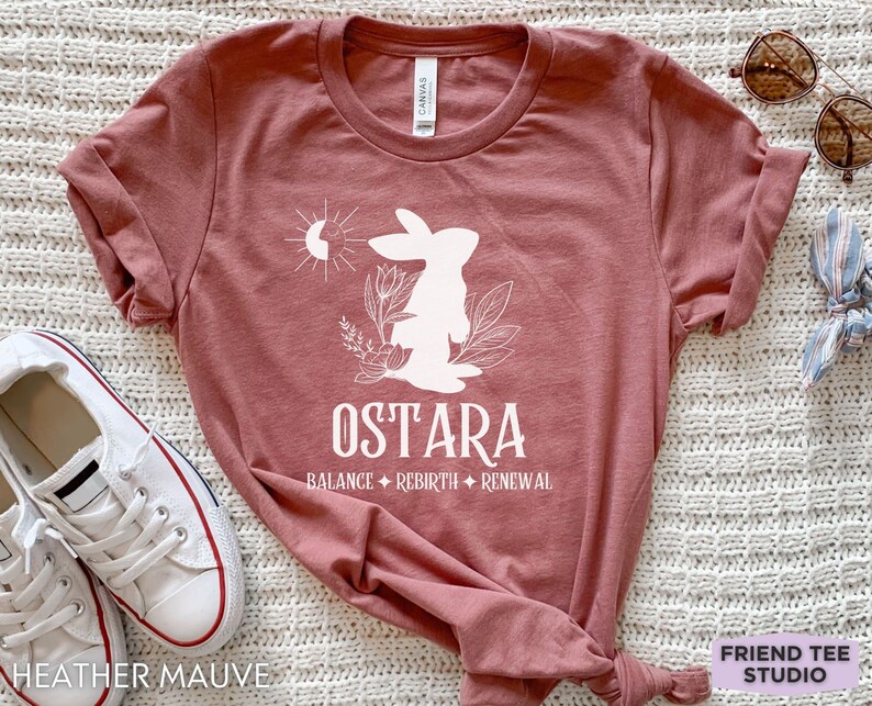 Puede incluir: Una camiseta de manga corta en color malva jaspeado con un gr&aacute;fico blanco de un conejo, flores y un s&iacute;mbolo de sol y luna. El texto dice OSTARA, seguido de las palabras BALANCE, REBIRTH y RENEWAL separadas por peque&ntilde;as estrellas de cuatro puntas.
