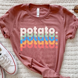 Puede incluir: Una camiseta de color rosa polvoriento con la palabra "potato" apilada verticalmente en una fuente retro. La palabra está superpuesta en varios colores, incluyendo blanco, verde, azul, naranja y rojo. La camiseta tiene cuello redondo y mangas cortas.