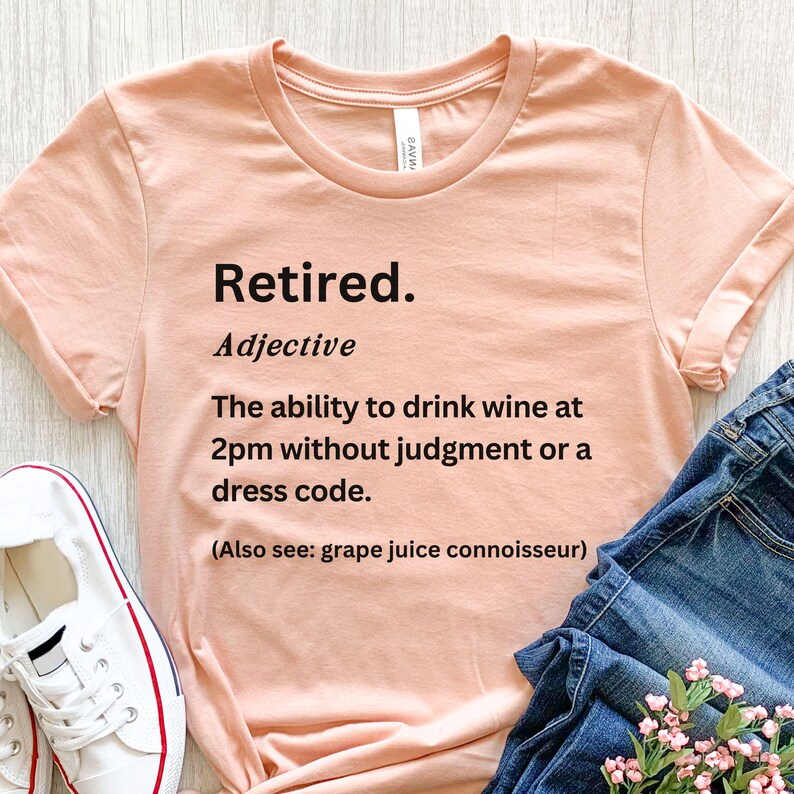 May include: A pale pink t-shirt with the word "Retired" printed in black. The definition below reads: "The ability to drink wine at 2pm without judgement or a dress code." Also includes "grape juice connoisseur".