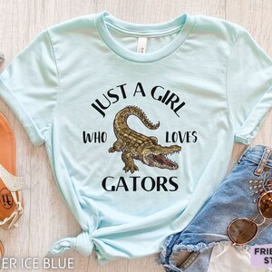 Puede incluir: Camiseta azul claro con el texto "JUST A GIRL WHO LOVES GATORS" y una ilustraci&oacute;n de un caim&aacute;n. La camiseta se muestra con pantalones cortos de mezclilla y gafas de sol.