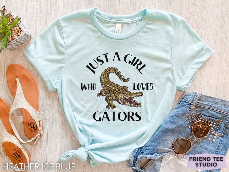Puede incluir: Camiseta azul claro con el texto "JUST A GIRL WHO LOVES GATORS" y una ilustraci&oacute;n de un caim&aacute;n. La camiseta se muestra con pantalones cortos de mezclilla y gafas de sol.