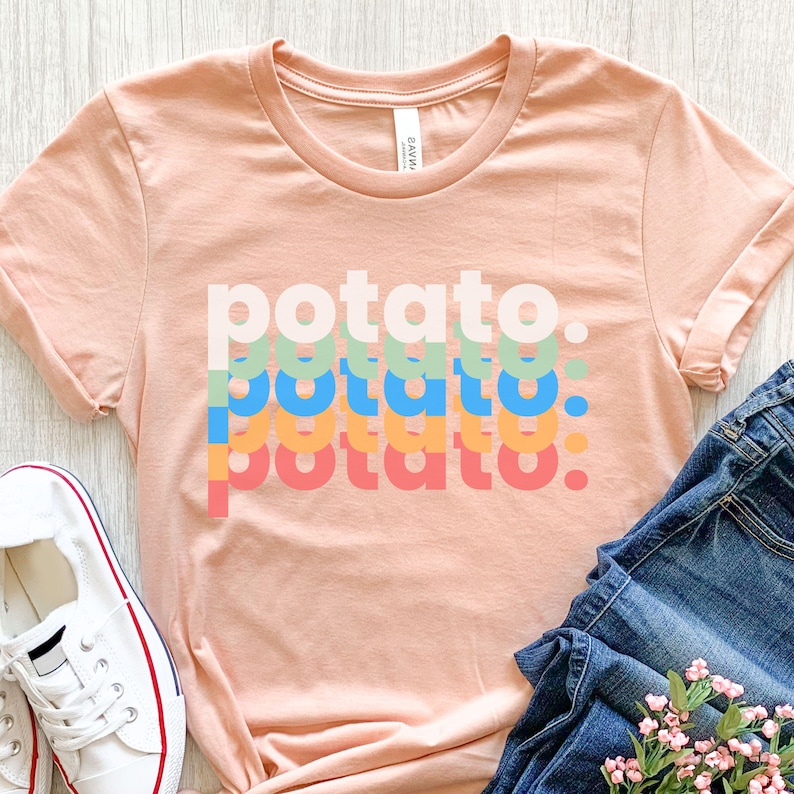 May include: A peach-colored short-sleeve t-shirt featuring the word potato printed four times in a stacked, overlapping design. The text appears in four distinct colors: light green, bright blue, orange, and coral, each with a period at the end.