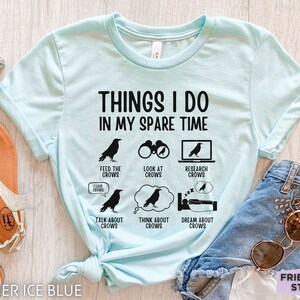 K&ouml;nnte beinhalten: Ein hellblaues T-Shirt mit dem Text "Things I do in my spare time" und einer Reihe von schwarzen Kr&auml;henillustrationen mit Text, der kr&auml;henbezogene Aktivit&auml;ten beschreibt, wie z. B. "Kr&auml;hen f&uuml;ttern", "Kr&auml;hen beobachten", "Kr&auml;hen recherchieren", "&Uuml;ber Kr&auml;hen sprechen", "An Kr&auml;hen denken" und "Von Kr&auml;hen tr&auml;umen".