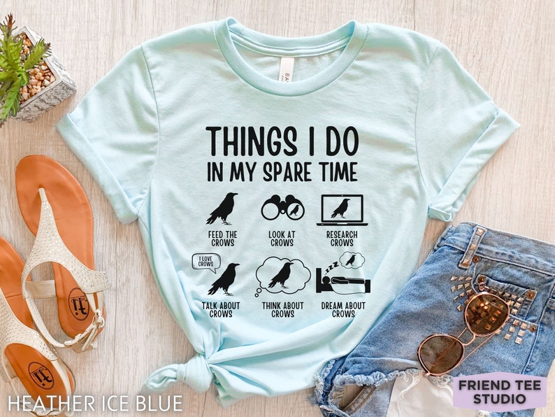 K&ouml;nnte beinhalten: Ein hellblaues T-Shirt mit dem Text "Things I do in my spare time" und einer Reihe von schwarzen Kr&auml;henillustrationen mit Text, der kr&auml;henbezogene Aktivit&auml;ten beschreibt, wie z. B. "Kr&auml;hen f&uuml;ttern", "Kr&auml;hen beobachten", "Kr&auml;hen recherchieren", "&Uuml;ber Kr&auml;hen sprechen", "An Kr&auml;hen denken" und "Von Kr&auml;hen tr&auml;umen".