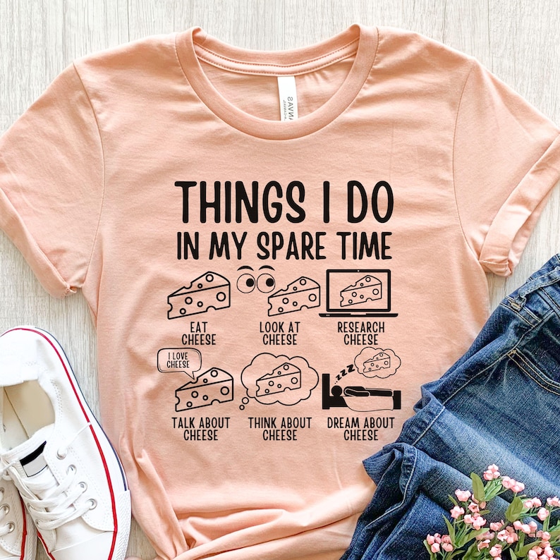 Puede incluir: Una camiseta de color melocot&oacute;n con un gr&aacute;fico negro que dice "Things I do in my spare time" y enumera actividades relacionadas con el queso. El gr&aacute;fico incluye ilustraciones de cu&ntilde;as de queso, una pantalla de computadora y una persona durmiendo.