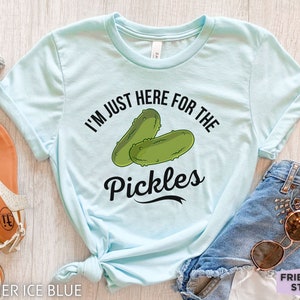 May include: Heather ice blue t-shirt with the text "I'm just here for the pickles" and a graphic of two green pickles. The shirt is paired with denim shorts and sunglasses.