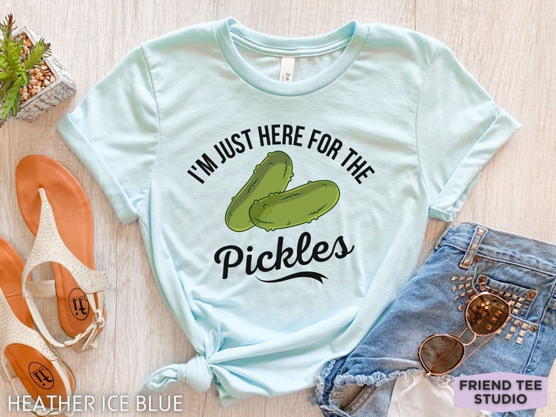 May include: Heather ice blue t-shirt with the text "I'm just here for the pickles" and a graphic of two green pickles. The shirt is paired with denim shorts and sunglasses.