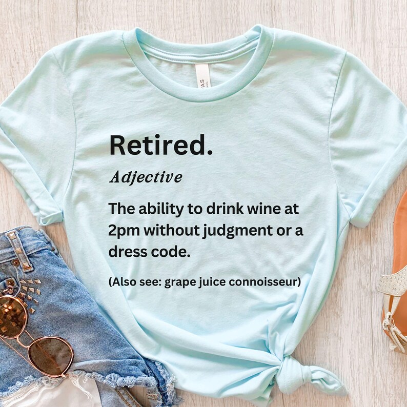 May include: Pale blue t-shirt featuring the word "Retired" and a definition printed in black. The definition humorously describes the freedom to drink wine at 2 PM without judgment. The shirt also mentions "grape juice connoisseur".