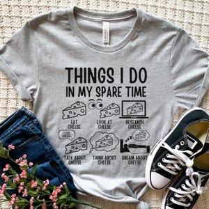 Puede incluir: Una camiseta gris con el texto "Things I do in my spare time" y una lista de actividades relacionadas con el queso: comer queso, mirar queso, investigar queso, hablar de queso, pensar en queso y so&ntilde;ar con queso.