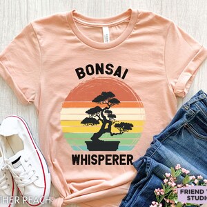 May include: A peach-colored t-shirt with a vintage-style graphic of a bonsai tree and the text "Bonsai Whisperer".
