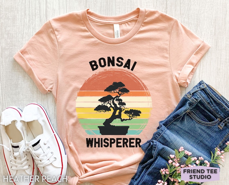 May include: A peach-colored t-shirt with a vintage-style graphic of a bonsai tree and the text "Bonsai Whisperer".
