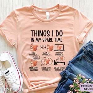 May include: A peach-colored t-shirt with the text "THINGS I DO IN MY SPARE TIME" and a series of illustrations of squirrels with text describing activities related to squirrels. The text includes "FEED SQUIRRELS", "LOOK AT SQUIRRELS", "RESEARCH SQUIRRELS", "TALK ABOUT SQUIRRELS", "THINK ABOUT SQUIRRELS", and "DREAM ABOUT SQUIRRELS".