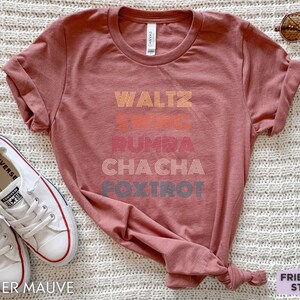 May include: Heather mauve t-shirt with the words "Waltz, Swing, Rumba, Cha Cha, Foxtrot" in a retro font. The shirt is styled with white sneakers and sunglasses. The text "Friend Tee Studio" is in the bottom right corner.