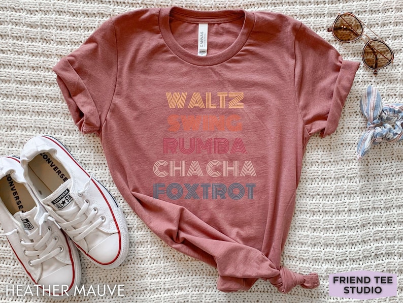 May include: Heather mauve t-shirt with the words "Waltz, Swing, Rumba, Cha Cha, Foxtrot" in a retro font. The shirt is styled with white sneakers and sunglasses. The text "Friend Tee Studio" is in the bottom right corner.