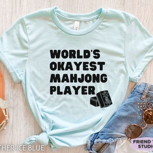 May include: A light blue t-shirt with black text that reads "World's Okayest Mahjong Player". The shirt is shown on a wooden surface with a pair of sandals and a pair of denim shorts with sunglasses.