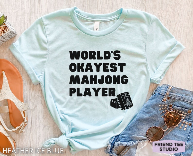 May include: A light blue t-shirt with black text that reads "World's Okayest Mahjong Player". The shirt is shown on a wooden surface with a pair of sandals and a pair of denim shorts with sunglasses.