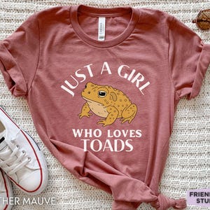 May include: Heather mauve t-shirt with the text "JUST A GIRL WHO LOVES TOADS" and a cartoon toad graphic. The shirt is paired with white sneakers. A pair of sunglasses and a hair tie are also visible.