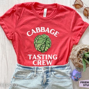 May include: A heather red short-sleeved t-shirt featuring a graphic of a green cabbage in the centre. The white text above the cabbage reads CABBAGE and the text below reads TASTING CREW. The shirt has a crew neckline and rolled sleeves.