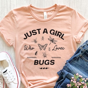 May include: A peach-colored t-shirt with the text "JUST A GIRL Who Loves BUGS" in black. The shirt features illustrations of various insects, including a butterfly, dragonfly, and beetles. White sneakers and blue jeans are visible.