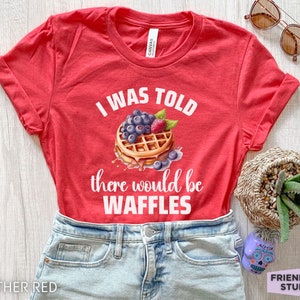 May include: Heather red t-shirt with the text "I WAS TOLD there would be WAFFLES" and a waffle graphic topped with blueberries and a raspberry. The shirt is displayed on a light wood surface.