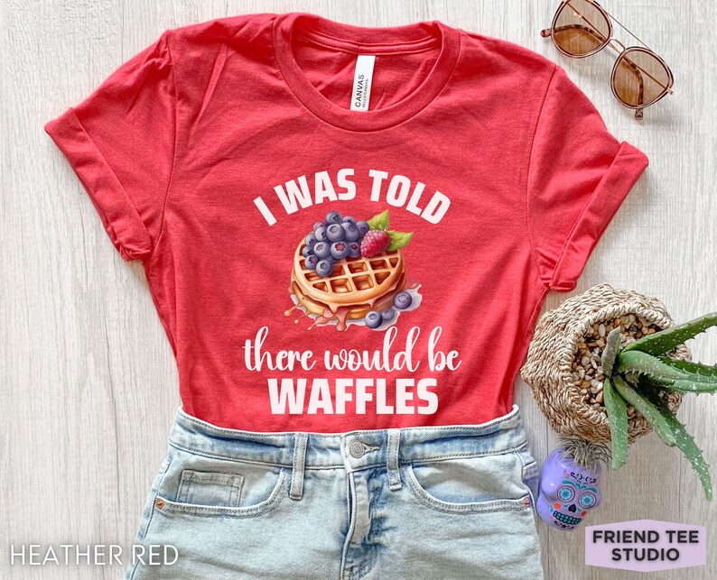 May include: Heather red t-shirt with the text "I WAS TOLD there would be WAFFLES" and a waffle graphic topped with blueberries and a raspberry. The shirt is displayed on a light wood surface.