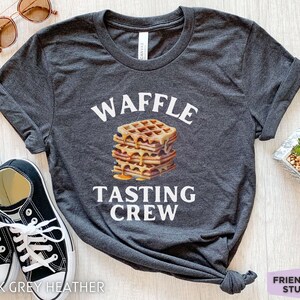 May include: Dark grey heather t-shirt with the words "WAFFLE TASTING CREW" in white. A stack of waffles with syrup is in the center. Black sneakers and sunglasses are in the background.