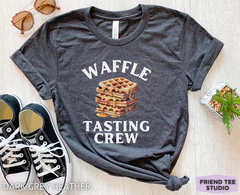 May include: Dark grey heather t-shirt with the words "WAFFLE TASTING CREW" in white. A stack of waffles with syrup is in the center. Black sneakers and sunglasses are in the background.