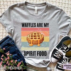 Pu&ograve; includere: Una maglietta grigio chiaro con la scritta "WAFFLES ARE MY SPIRIT FOOD" sopra una pila di waffle con sciroppo. Il design presenta una combinazione di colori retr&ograve; con strisce arancioni, rosa, gialle e blu. La maglietta &egrave; abbinata a jeans blu e scarpe da ginnastica nere.