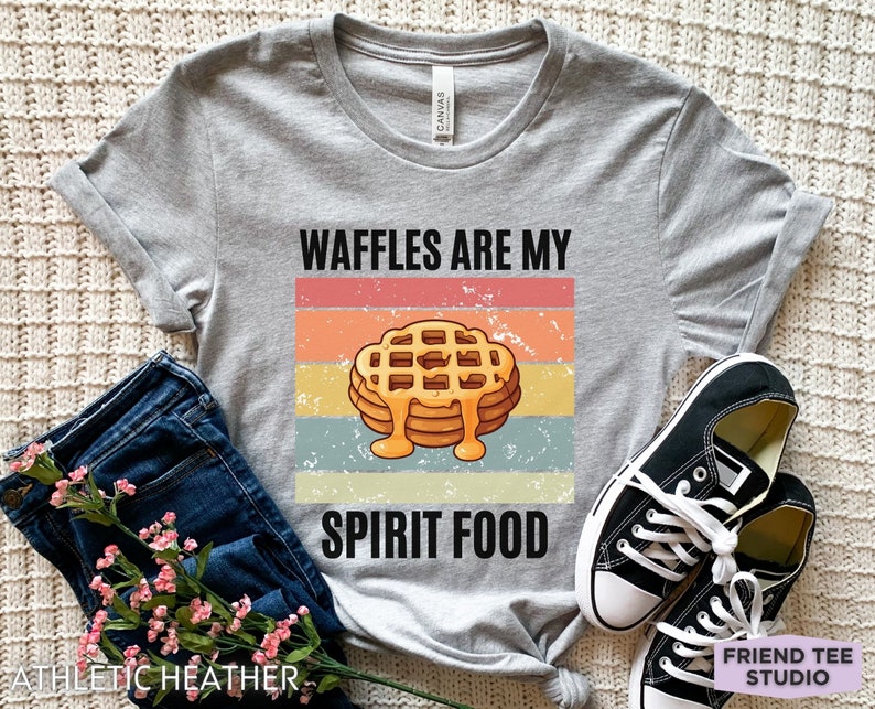 Pu&ograve; includere: Una maglietta grigio chiaro con la scritta "WAFFLES ARE MY SPIRIT FOOD" sopra una pila di waffle con sciroppo. Il design presenta una combinazione di colori retr&ograve; con strisce arancioni, rosa, gialle e blu. La maglietta &egrave; abbinata a jeans blu e scarpe da ginnastica nere.