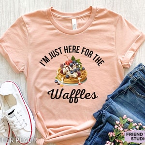 Op de afbeelding: Een perzikkleurig T-shirt met de tekst "I'm just here for the waffles" in zwart. Een wafel met slagroom, fruit en chocoladesiroop is afgebeeld boven de tekst.