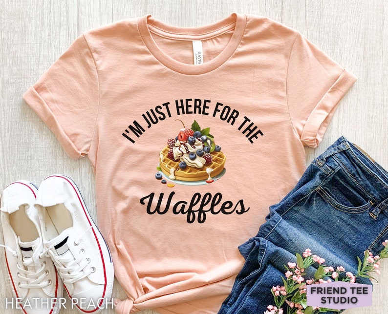 Op de afbeelding: Een perzikkleurig T-shirt met de tekst "I'm just here for the waffles" in zwart. Een wafel met slagroom, fruit en chocoladesiroop is afgebeeld boven de tekst.