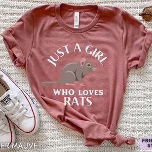 May include: A heather mauve short-sleeve t-shirt featuring a graphic of a brown rat in the center. The white text above and below the rat reads, "JUST A GIRL WHO LOVES RATS." The shirt has a crew neckline and is displayed with white canvas sneakers.