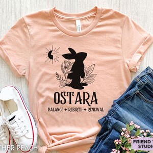 Puede incluir: Una camiseta de manga corta color melocot&oacute;n jaspeado con un gr&aacute;fico negro de un conejo, flores y un sol. El texto dice OSTARA, BALANCE, REBIRTH, RENEWAL. La camiseta se muestra con zapatillas blancas y pantalones vaqueros azules.