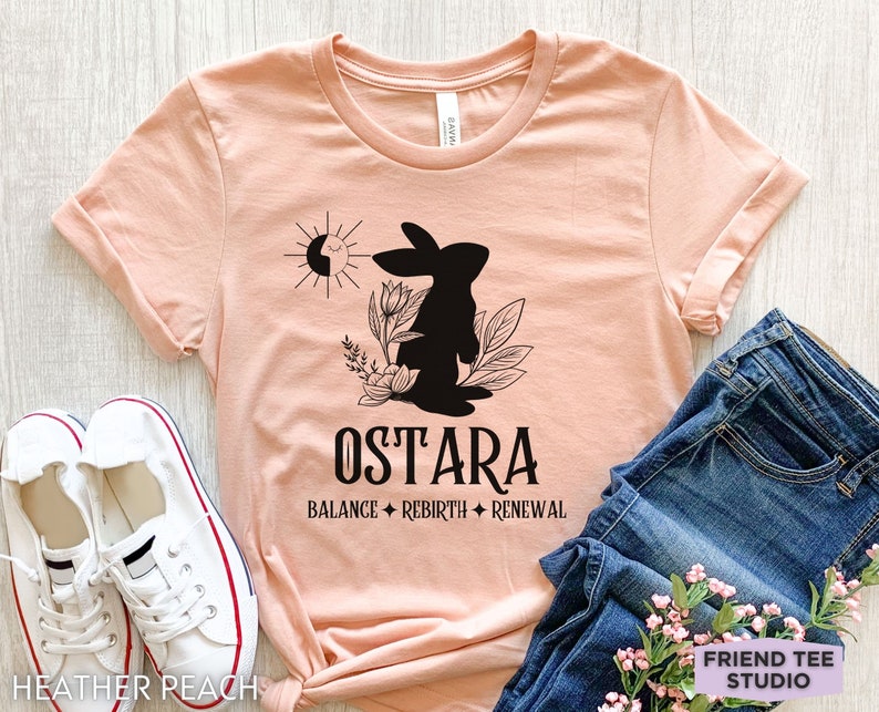 Puede incluir: Una camiseta de manga corta color melocot&oacute;n jaspeado con un gr&aacute;fico negro de un conejo, flores y un sol. El texto dice OSTARA, BALANCE, REBIRTH, RENEWAL. La camiseta se muestra con zapatillas blancas y pantalones vaqueros azules.