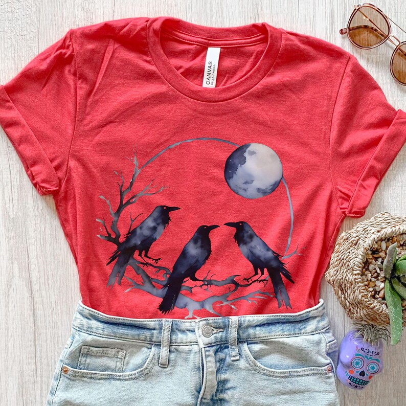 May include: Red t-shirt featuring a watercolor design of three black crows perched on a branch, with a full moon in the background. The shirt is made of a soft material and has a classic crew neck.
