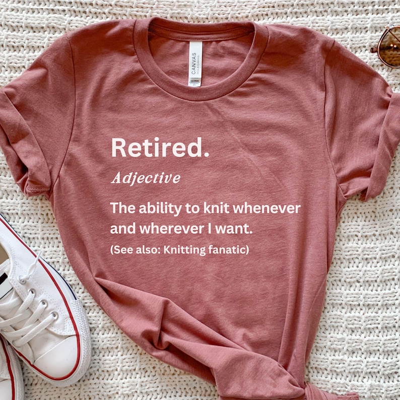 Puede incluir: Camiseta rosa polvoriento con la palabra "Retired" en letras blancas. Debajo, la definici&oacute;n dice "La capacidad de tejer cuando y donde quiera." La camiseta tambi&eacute;n incluye "(Ver tambi&eacute;n: Knitting fanatic)."